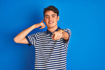 Naklejka premium Teenager boy wearing casual t-shirt standing over blue isolated background smiling doing talking on the telephone gesture and pointing to you. Call me.
