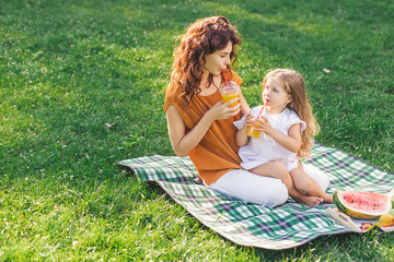 mother and daughter drinking orange juice from plastic cups sitting on the blanket in the park