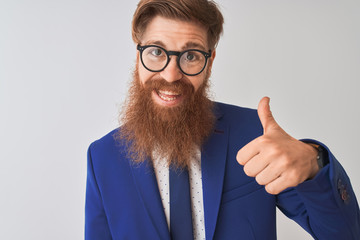 Young redhead irish businessman wearing suit and glasses over isolated white background doing happy thumbs up gesture with hand. Approving expression looking at the camera with showing success.