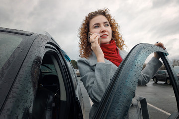 A business woman is standing near her car in a parking lot and talking on her cell phone. Beautiful young woman driving on a cloudy day