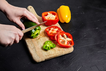 Pepper slicing. Hands, knife and red, green, yellow bell peppers, wooden cutting board on stone table. Sliced sweet peppers on black background. Vegetable ingredient, cooking diet salad, healthy food
