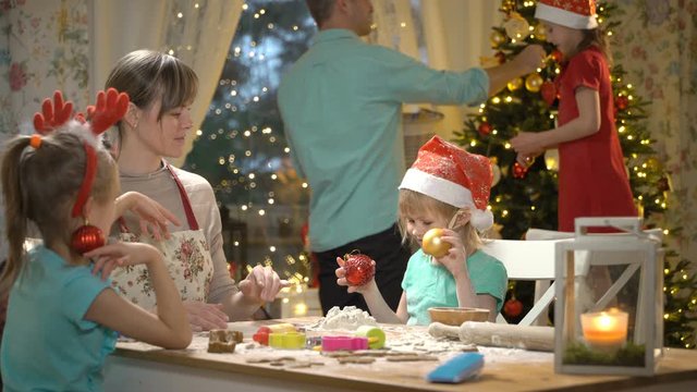Children with their mom making gingerbread cookies for family dinner on Christmas eve in dining room. Father with little daughter decorating Christmas tree. Children laughing and fooling around