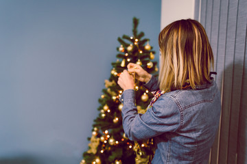 Back view of woman female wearing blue shirt decorating Christmas tree at home having fun for holidays new year
