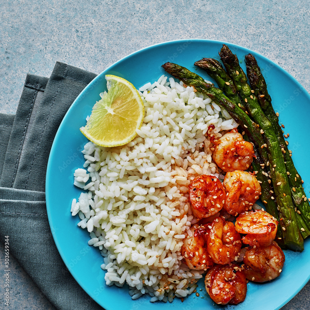 Wall mural Teriyaki coated shrimps stir fry with grilled asparagus, basmati rice and sesame seeds served on a blue plate. Easy delicious meal. Top view, directly above shot.