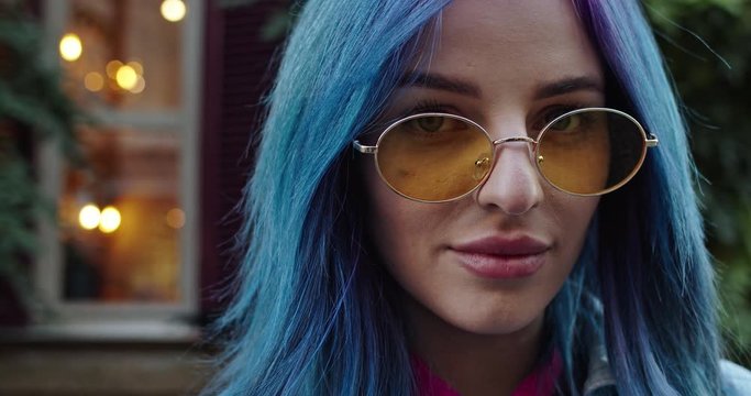 Close up of the face of the young pretty Caucasian hipster girl with blue hair and in sunglasses looking straight to the camera and smiling happily. Outdoor. Portrait.