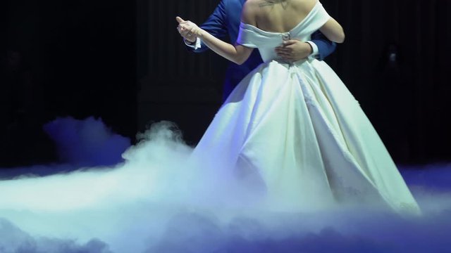 Happy beautiful newlyweds dancing in the restaurant in puffs of fluffy smoke on the floor close up. The bride and groom in their first wedding dance on black isolated background. Spotlights shine.