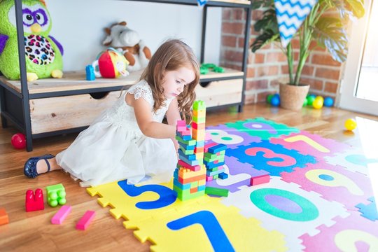 Adorable blonde toddler playing with building blocks toy around lots of toys at kindergarten