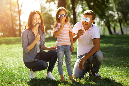 Family Of Three Using Funny Paper Masks On The Sticks Outdoors And Looking At The Camera