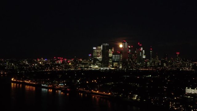 Aerial drone distant view moon rising over Canary Wharf London's Docklands Isle of Dogs and Thames River at night 