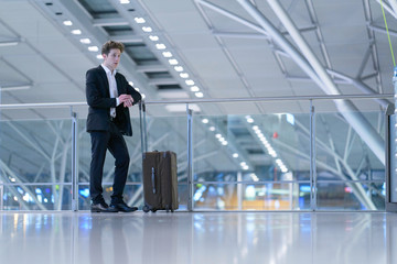 Young relaxed smiling businessman standing with his rolling suitcase in the airport, leaning with his arm on a guard railing