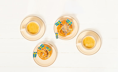 Swedish cinnamon buns (kanelbullar) and two cups of coffee on the white wooden table. Swedish flags. Coffee break (fika) concept. Top view.