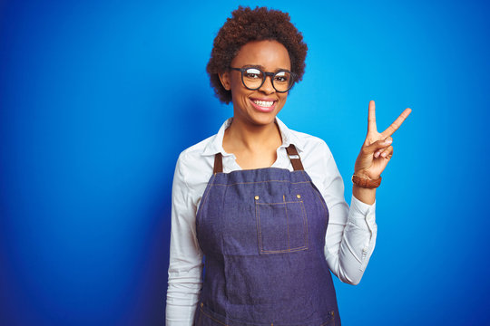 Young african american woman shop owner wearing business apron over blue background smiling with happy face winking at the camera doing victory sign. Number two.