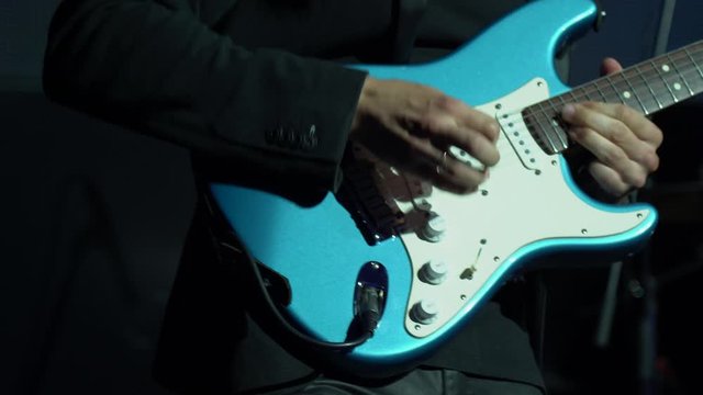 Handsome white man guitar player in a black jacket quickly and actively play with pleasure on a blue and white professional electric guitar on a black background in the light of spotlights close up.