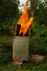 Parts of an old wooden structure burn in an iron barrel. There is a high flame. Apple trees with unripe fruits and tall grass are visible in the background.