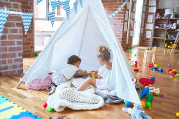 Fototapeta na wymiar Beautiful toddlers playing inside tipi over blanket around lots of toys at kindergarten
