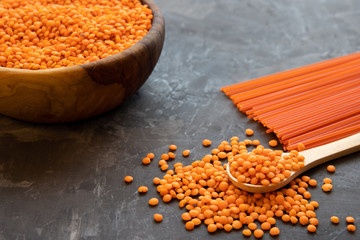 A round wooden bowl full of red lentils stands on a dark table. Nearby lies a wooden spoon and spaghetti, pasta from orange lentils