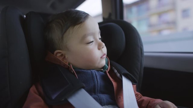 Medium shot of sleepy toddler boy sitting in car seat of moving car and looking out window