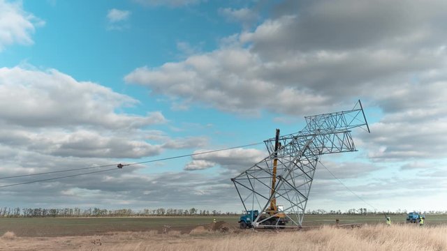 The crane lifts the electric support of the new power line, workers direct the direction in the field against a blue sky covered with clouds, a distant view. Time Laps video of a power line