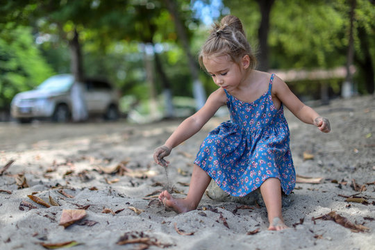 Little girl in a blue dress plays with sand on the beach sprinkling sand on her foot.