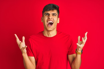 Young indian man wearing t-shirt standing over isolated red background crazy and mad shouting and yelling with aggressive expression and arms raised. Frustration concept.