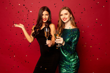  two beautiful models, a redhead and a brunette in New Year's dresses, having fun and smiling with glasses of champagne, confetti flying around on a red background. New Year or Christmas photo - obrazy, fototapety, plakaty