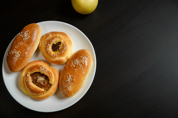 Delicious sesame buns or pies on white plate and yellow apple on black wooden table background. Tasty unhealthy snack for lunch. Overweight, diet, nutrition, eating habits concept. Place for text