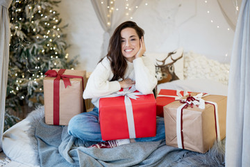 Obraz na płótnie Canvas Young beautiful brunette woman wearing christmas sweater sitting on the sofa with wrapped presents in room decorated for celebrating new year and christmas festive mood