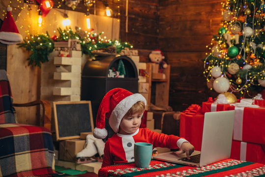 Santa helper baby boy with laptop. Bright New Years interior. Xmas tree. Indoor. Child at Christmas tree and fireplace on Xmas eve. Christmas time. Christmas attributes.
