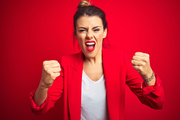 Young beautiful business woman standing over red isolated background angry and mad raising fists frustrated and furious while shouting with anger. Rage and aggressive concept.