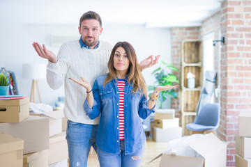 Young beautiful couple standing at new home around cardboard boxes clueless and confused expression...