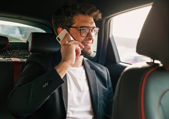 Young and attractive man with a beard and glasses speaks with his smartphone smiling inside a car. Business