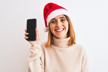 Beautiful redhead woman wearing christmas hat showing smartphone screen with a happy face standing and smiling with a confident smile showing teeth