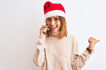 Beautiful redhead woman wearing christmas hat talking on smartphone pointing and showing with thumb up to the side with happy face smiling