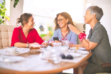 Meeting of middle age women having lunch and drinking coffee. Mature friends smiling happy using smartphone at home on a sunny day