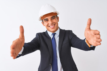 Young handsome architect man wearing suit and helmet over isolated white background looking at the camera smiling with open arms for hug. Cheerful expression embracing happiness.