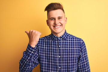 Fototapeta premium Young handsome man wearing casual shirt standing over isolated yellow background smiling with happy face looking and pointing to the side with thumb up.