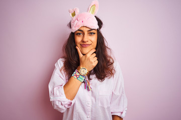 Young beautiful woman wearing pajama and sleep mask over isolated pink background looking confident at the camera with smile with crossed arms and hand raised on chin. Thinking positive.