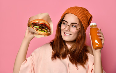 Beautiful teenage girl with red hair and hat holding burger and beverage in both hands. - obrazy, fototapety, plakaty