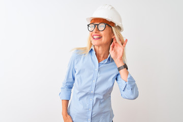 Middle age architect woman wearing glasses and helmet over isolated white background smiling with hand over ear listening an hearing to rumor or gossip. Deafness concept.