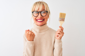 Middle age painter woman holding paint brush standing over isolated white background screaming proud and celebrating victory and success very excited, cheering emotion