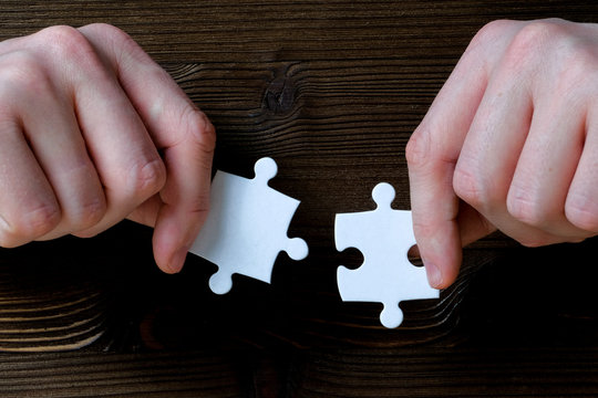 Hands Hold Two Pieces Of A White Puzzle On A Wooden Background.