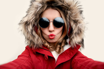 Close up portrait of attractive woman blowing red lips sending sweet air kiss stretching hand for taking selfie wearing red jacket with fur hood