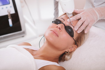 Close up of a mature woman  getting her eyebrow tattoo removed at cosmetology clinic. Dermatologist removing tattooed eyebrows on a female client using laser - obrazy, fototapety, plakaty
