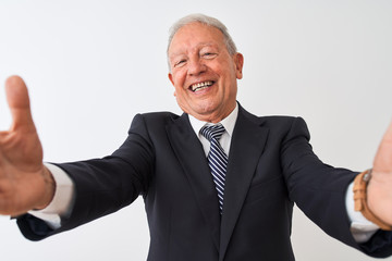 Senior grey-haired businessman wearing suit standing over isolated white background looking at the camera smiling with open arms for hug. Cheerful expression embracing happiness.
