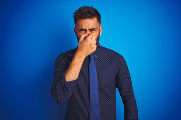 Young indian businessman wearing elegant shirt and tie standing over isolated blue background smelling something stinky and disgusting, intolerable smell, holding breath with fingers on nose. 