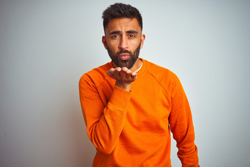 Fototapeta premium Young indian man wearing orange sweater over isolated white background looking at the camera blowing a kiss with hand on air being lovely and sexy. Love expression.