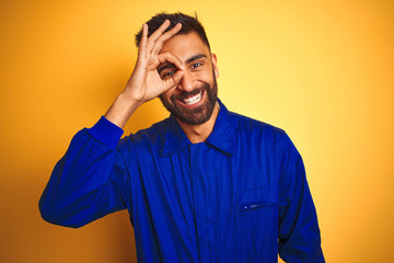 Handsome indian worker man wearing uniform over isolated yellow background doing ok gesture with hand smiling, eye looking through fingers with happy face.