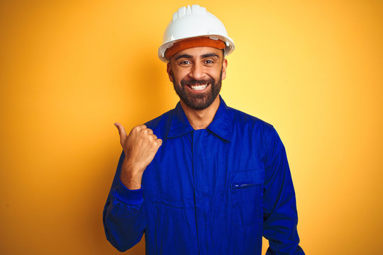 Handsome indian worker man wearing uniform and helmet over isolated yellow background smiling with happy face looking and pointing to the side with thumb up.