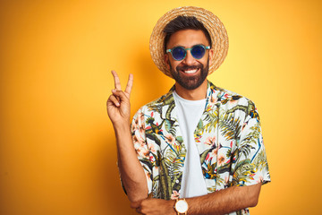Indian man on vacation wearing floral shirt hat sunglasses over isolated yellow background smiling...