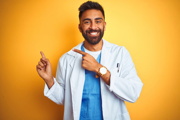 Young indian doctor man standing over isolated yellow background smiling and looking at the camera...